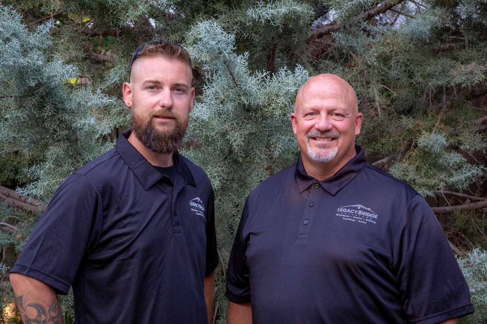Britt and Eric, core leaders at Legacy Ridge Exteriors, stand side by side smiling in matching black polo shirts with company logos, framed by natural evergreen foliage.