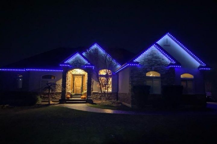 Luxurious stone-and-stucco residence glowing with vivid blue LED lights tracing rooflines and gables, creating a modern, icy holiday ambiance against dark night.