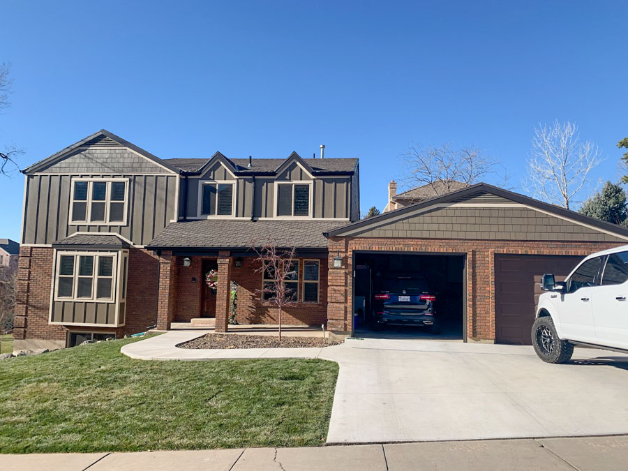 Elegant two-story home with dark gray horizontal siding, brick lower level, open garage revealing a car, and festive Christmas lights on the front porch.