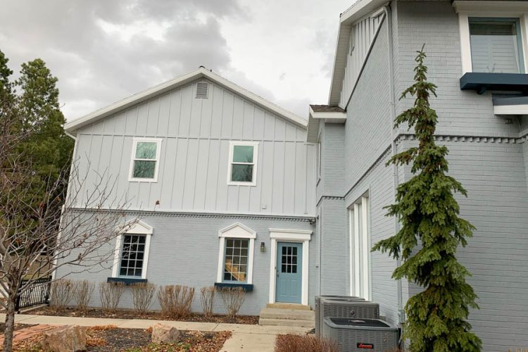 Charming farmhouse-style residence with light gray vertical siding, white-trimmed windows, a welcoming blue front door, and an evergreen tree in the front yard.