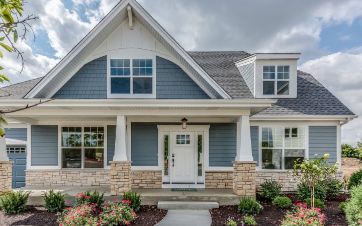 Beautiful modern farmhouse-style home with blue shingle and siding accents, large multi-pane windows, white trim, stone foundation, and colorful flowerbeds under a dramatic sky.