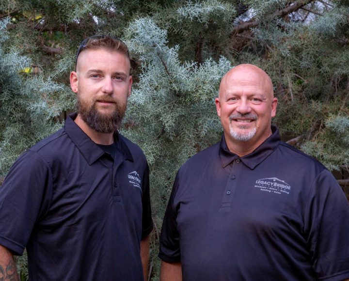Britt and Eric, core leaders at Legacy Ridge Exteriors, stand side by side smiling in matching black polo shirts with company logos, framed by natural evergreen foliage.