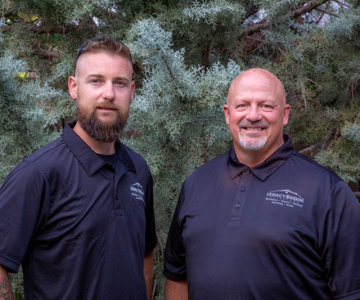 Britt and Eric, core leaders at Legacy Ridge Exteriors, stand side by side smiling in matching black polo shirts with company logos, framed by natural evergreen foliage.