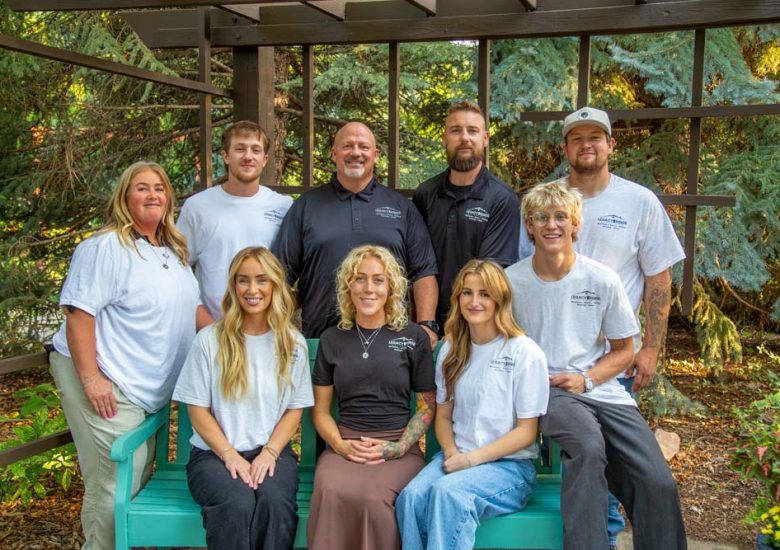 The full Legacy Ridge Exteriors team poses together outdoors under a wooden pergola, wearing branded company shirts, with lush green trees and a turquoise bench in the background.