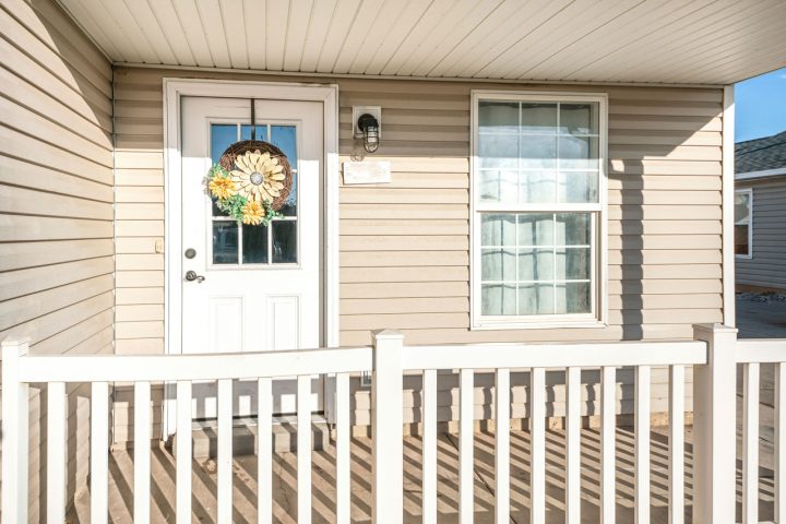 Cozy front porch featuring a white entry door adorned with a sunflower wreath, beige horizontal siding, white railings, and bright natural lighting under a clear sky.