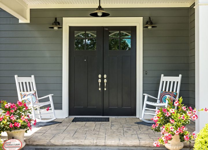 Inviting entryway with dark gray siding, black double doors with glass panels, white rocking chairs, and vibrant pink potted flowers on a stamped concrete porch.