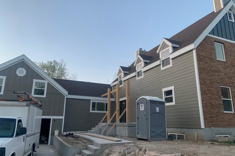 A modern home under construction featuring gray horizontal siding, white trim, and a temporary restroom on-site, with a work van parked nearby and exposed foundation steps.