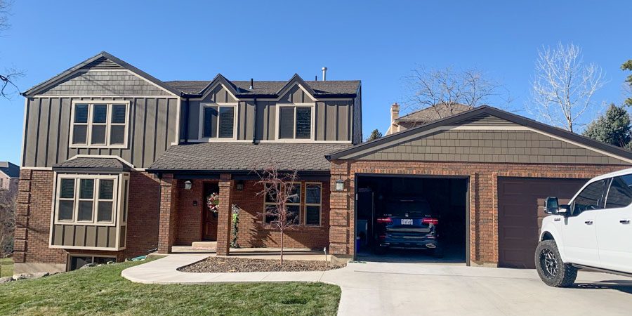 Elegant two-story home with dark gray horizontal siding, brick lower level, open garage revealing a car, and festive Christmas lights on the front porch.
