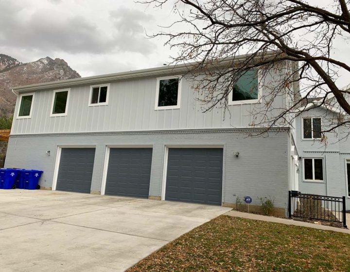 Two-story home with light blue horizontal siding, three dark gray garage doors, and a scenic mountain backdrop under cloudy skies.