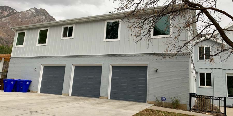 Two-story home with light blue horizontal siding, three dark gray garage doors, and a scenic mountain backdrop under cloudy skies.